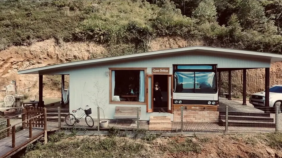 Fachada da Casa Ônibus: casa de madeira clara com a frente de um ônibus embutida, letreiro "Casa Ônibus", deck e bicicleta, com o morro de mata atrás