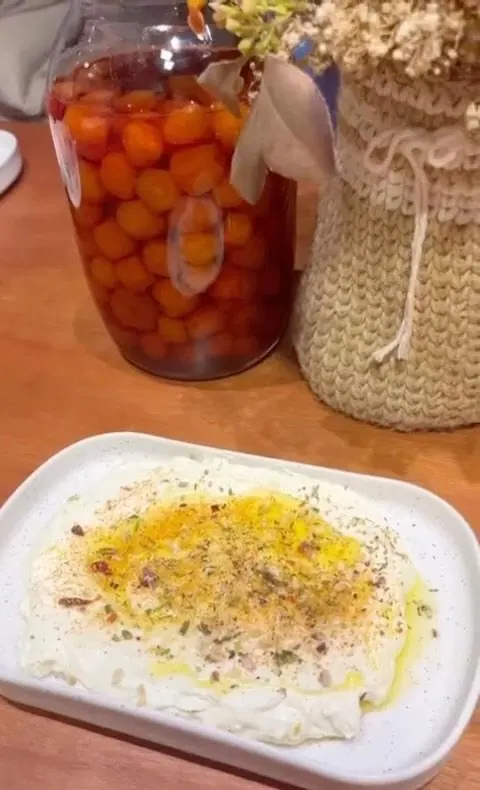 Travessa de coalhada com azeite e temperos, ao lado de um pote de conserva e um vaso de flores secas